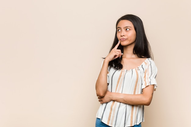 Young cute chinese teenager Young blonde woman wearing a coat against a pink background looking sideways with doubtful and skeptical expression.