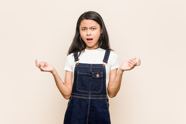 Young cute chinese teenager young blonde woman wearing a coat against a pink wall screaming with rage.