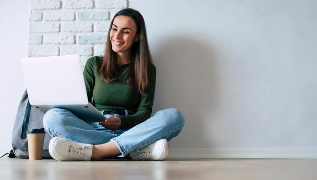 Young cute confident student woman in casual clothes is using a laptop and smiling while sitting on the floor on gray background Study education university college graduate concept