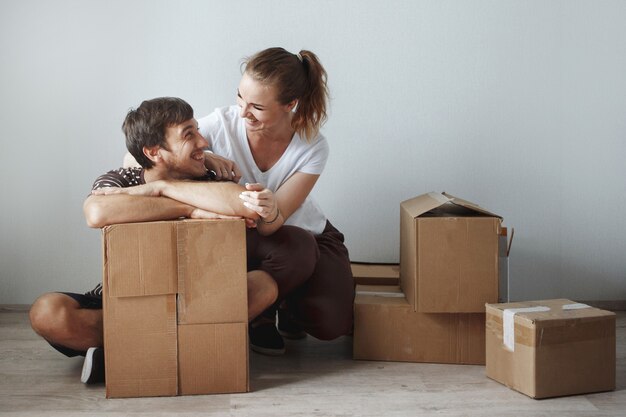Young cute couple just married sitting on floor in a new apartment after repair among the cardboard boxes are happy and smiling.