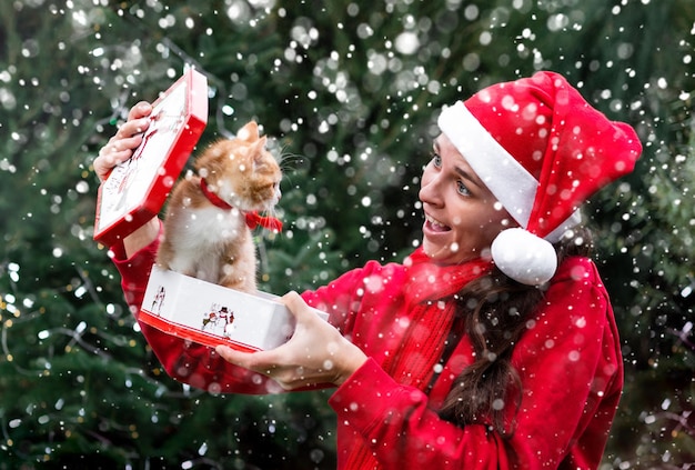 Young cute European woman in red Santa hat gets little red kitten as Christmas gift