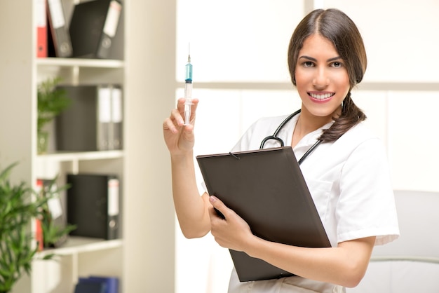 Young cute female doctor in a white coat, with a stethoscope over the neck holding an injection and folder, with a smile looking at the camera.