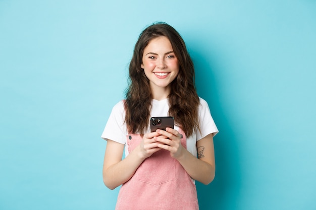 Молодая милая девушка держит мобильный телефон, улыбается и смотрит в камеру. Женщина, использующая приложение для смартфона, общение в социальных сетях или покупки в Интернете, синий фон