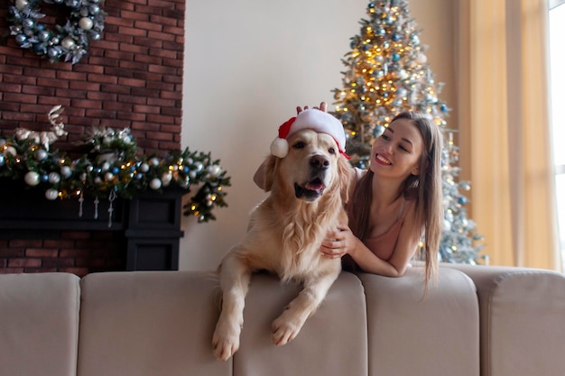 young cute girl hugging dog in santa hat at home near new year tree and looking at copy space