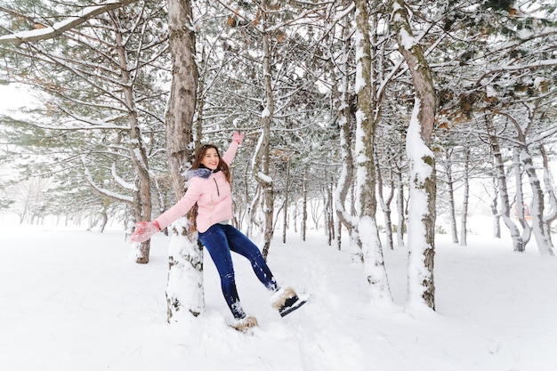 ピンクの暖かいジャケットとジーンズが木にもたれて雪の背景で楽しんで若いかわいい女の子。冬のエンターテイメント