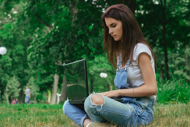 молодая милая девушка сидит на траве и печатает на ноутбуке