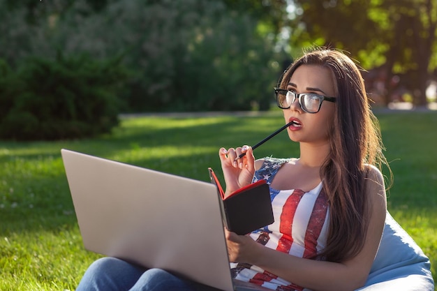 Young cute girl with laptop sit on bean bag in garden or park on green grass modern lifestyle concept