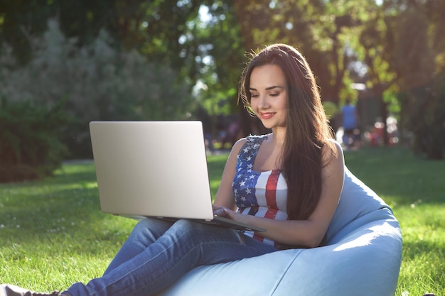 Young cute girl with laptop, sit on bean bag in garden or park, on green grass. Online shopping concept