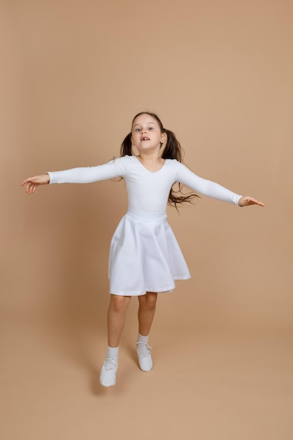 Young cute girl with long dark hair in white dress socks and gymnastics shoes whirling dancing spreading hands raising leg having fun on brown background