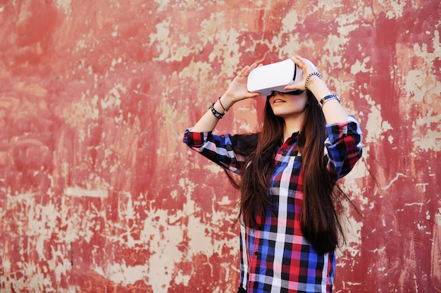 Young cute girl with long hair in plaid shirt in virtual reality glasses on red grunge wall background.