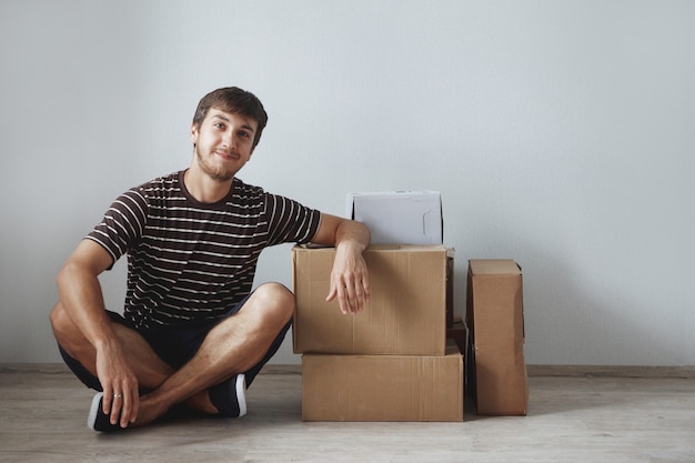 Young cute guy sits on the floor in a new apartment after repair among the cardboard boxes, happy and smiling.