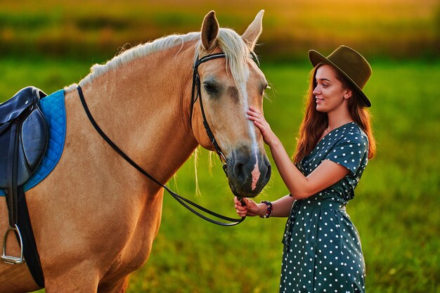 Giovane donna sorridente soddisfatta allegra felice sveglia che abbraccia e che accarezza il bellissimo cavallo palomino biondo al prato al tramonto