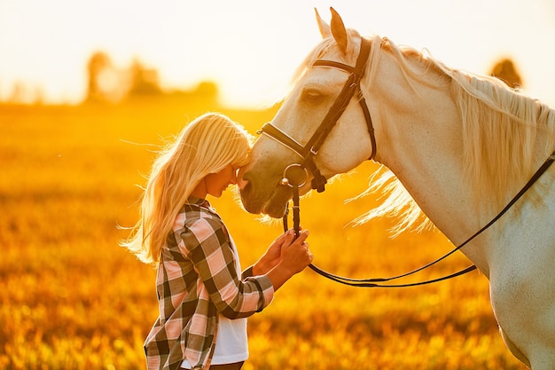 Giovane donna sorridente soddisfatta allegra felice sveglia che abbraccia e che accarezza il bellissimo cavallo bianco al prato al tramonto all'ora d'oro