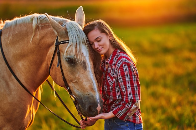Giovane donna sorridente soddisfatta allegra felice sveglia con gli occhi chiusi godendo di abbracciare e accarezzare il bellissimo cavallo palomino biondo al prato al tramonto