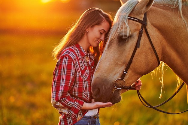 Молодая милая счастливая радостная довольная улыбающаяся женщина с закрытыми глазами, наслаждающаяся объятиями и поглаживанием красивой белокурой лошади Паломино на лугу на закате