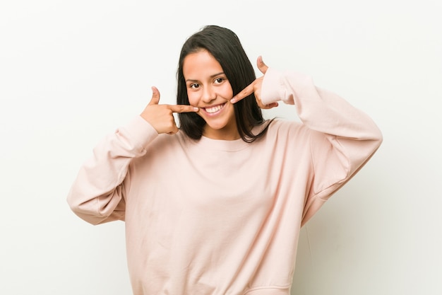 Young cute hispanic teenager woman smiles, pointing fingers at mouth.