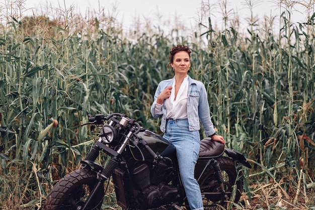 Young cute model in denim jacket sitting on black retro motorcycle. Beautiful woman and motorbike. Biker girl posing on bike at the background of grass. Biker concept