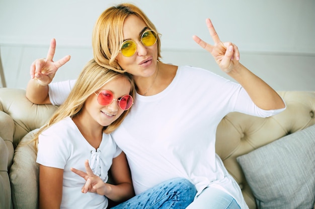 Young cute mother and little daughter in colorful sunglasses have a fun and hugging on the couch at home