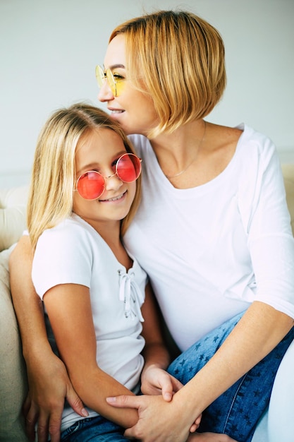 Young cute mother and little daughter in colorful sunglasses have a fun and hugging on the couch at home