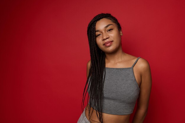 Young cute woman, attractive charming African American female athlete with fashionable stylish long hair and dreadlocks, in gray sportswear posing against red colored background with copy space for ad