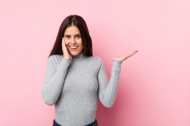 Young cute woman holds copy space on a palm, keep hand over cheek. Amazed and delighted.