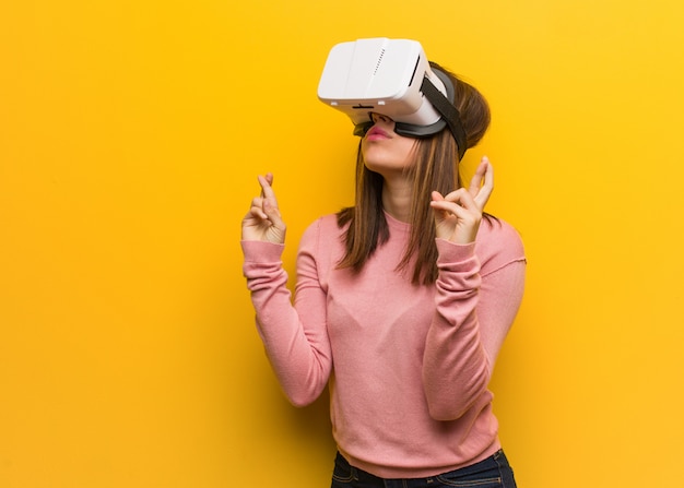 Young cute woman wearing a virtual reality goggles crossing fingers for having luck