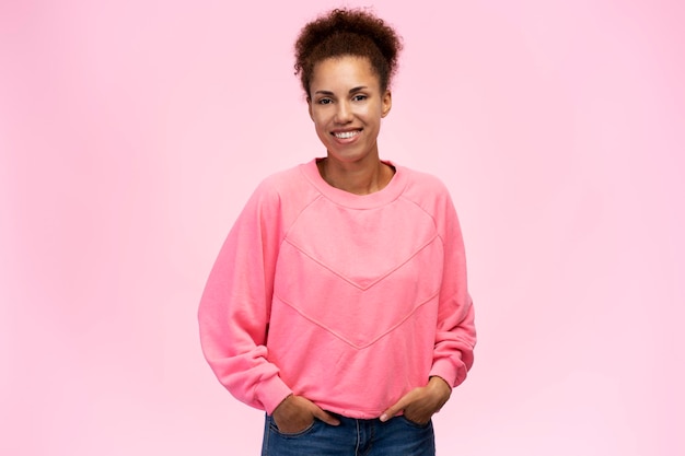 Young cute woman with afro hair holding hands on jeans pockets smiling at camera pink background