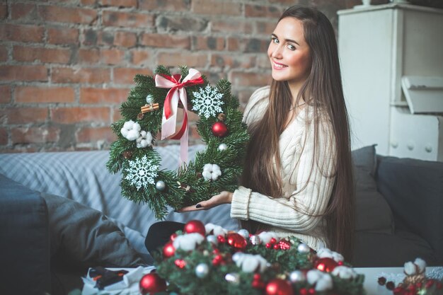 自宅で赤いガラスのボール、コーン、リボン、シナモンとクリスマス常緑樹の花輪を持つ若いかわいい女性