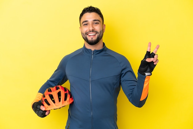 Young cyclist arab man isolated on yellow background smiling and showing victory sign