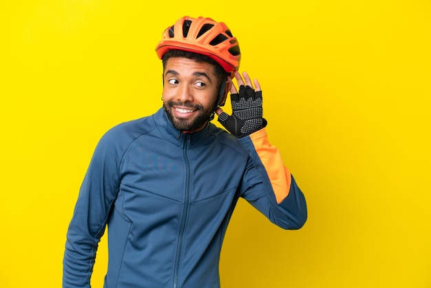 Young cyclist Brazilian man isolated on yellow background listening to something by putting hand on the ear