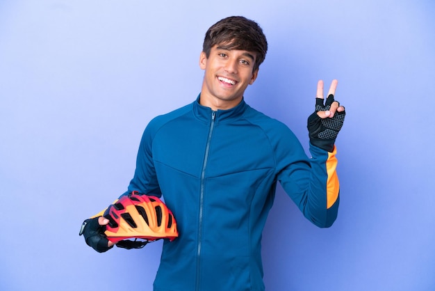 Young cyclist caucasian man isolated on purple background smiling and showing victory sign