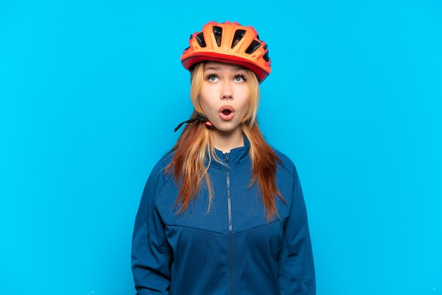 Young cyclist girl isolated on blue background looking up and with surprised expression