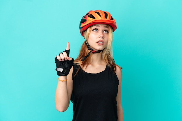 Молодой велосипедист девушка на изолированном синем фоне, указывая вверх и удивлен