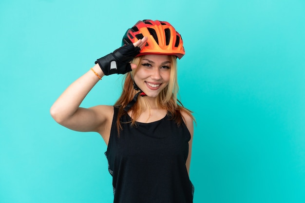 Молодая велосипедистка на изолированном синем фоне, салютуя рукой с счастливым выражением лица