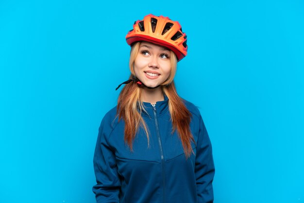 Young cyclist girl isolated on blue background thinking an idea while looking up