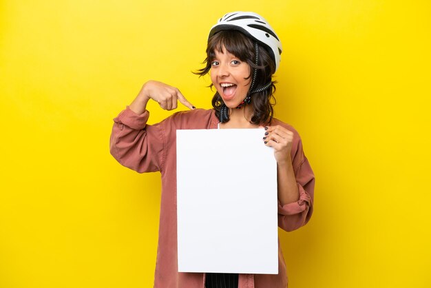 노란색 배경에 격리된 젊은 자전거 타는 라틴 여성이 행복한 표정으로 빈 현수막을 들고 가리키고 있습니다.