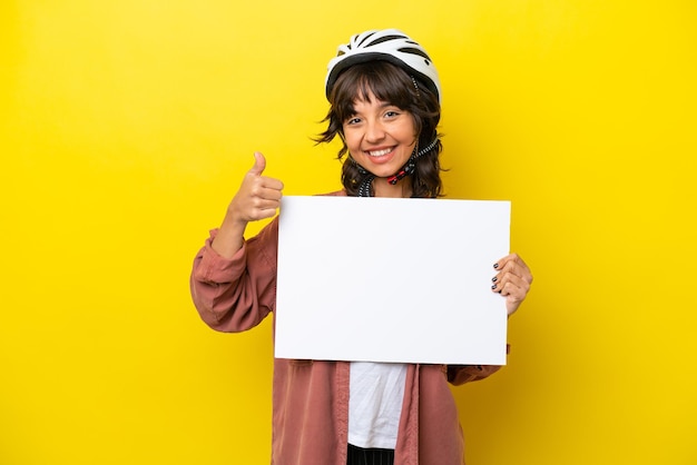 Молодая латинская женщина-велосипедист на желтом фоне держит пустой плакат с большим пальцем вверх