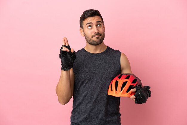 Young cyclist man isolated on pink background with fingers crossing and wishing the best