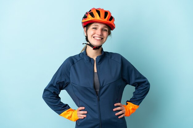 Young cyclist woman isolated on blue background posing with arms at hip and smiling