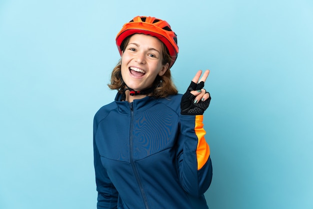 Young cyclist woman isolated on blue background smiling and showing victory sign