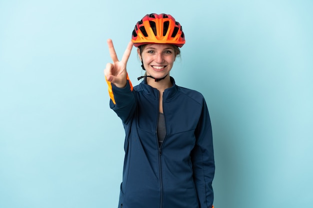 Young cyclist woman isolated on blue smiling and showing victory sign