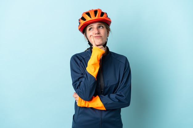 Young cyclist woman isolated on blue wall having doubts while looking up