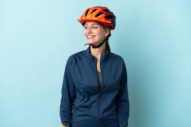 Young cyclist woman isolated on blue wall looking to the side and smiling