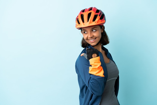 Young cyclist woman isolated on blue wall pointing to the side to present a product