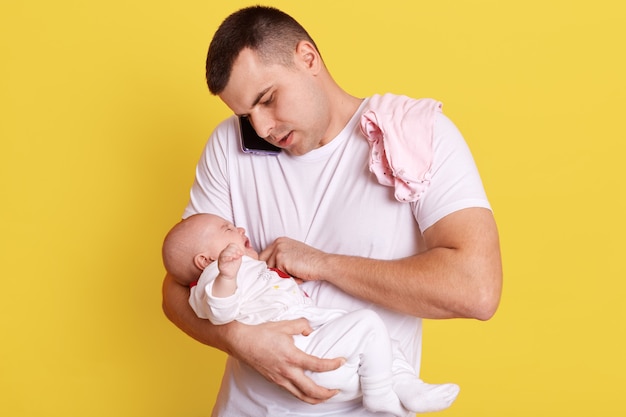 Young dad being at home with his baby son or girl, standing isolated over yellow wall, talking on phone with somebody and trying to put soother for newborn baby.