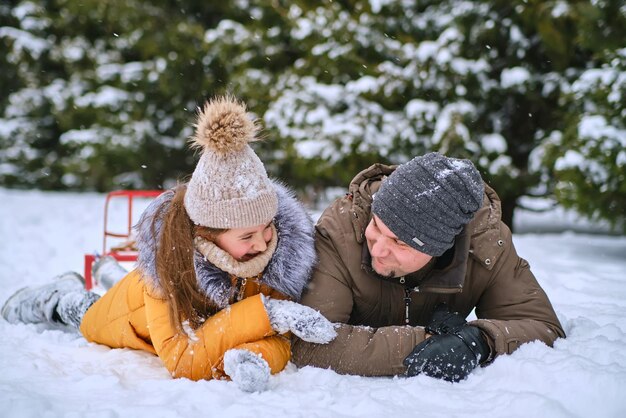 若いお父さんと彼の小さなかわいい娘は、雪の降る冬に屋外で楽しんでいます