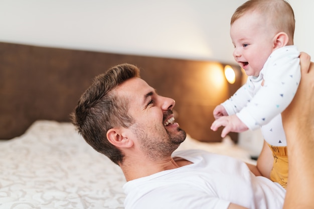 young dad lying in bed in bedroom and lifting his laughing baby boy