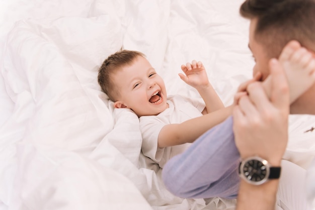 寝室の幸せな家族のベッドで自宅で彼の赤ちゃんの笑顔の息子と遊んで笑っている若いお父さん