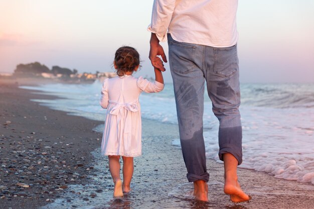 小さな娘と若いお父さんが夕日の海のビーチに沿って歩く