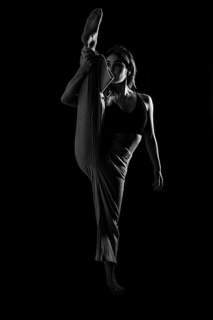 Молодая танцовщица в студийной фотосессии с балетом на черном фоне поднимает ногу